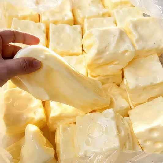A hand holding a pale yellow, cheese-shaped novelty item or soap, surrounded by many similar pale yellow cube-shaped pieces in a clear plastic bag.